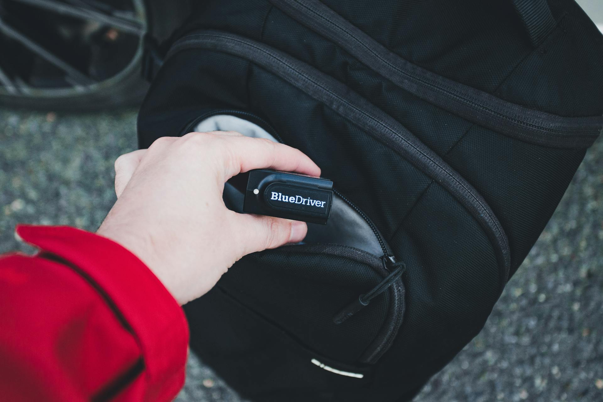 Close-up of hand holding BlueDriver scan tool near black backpack, emphasizing travel and technology use.