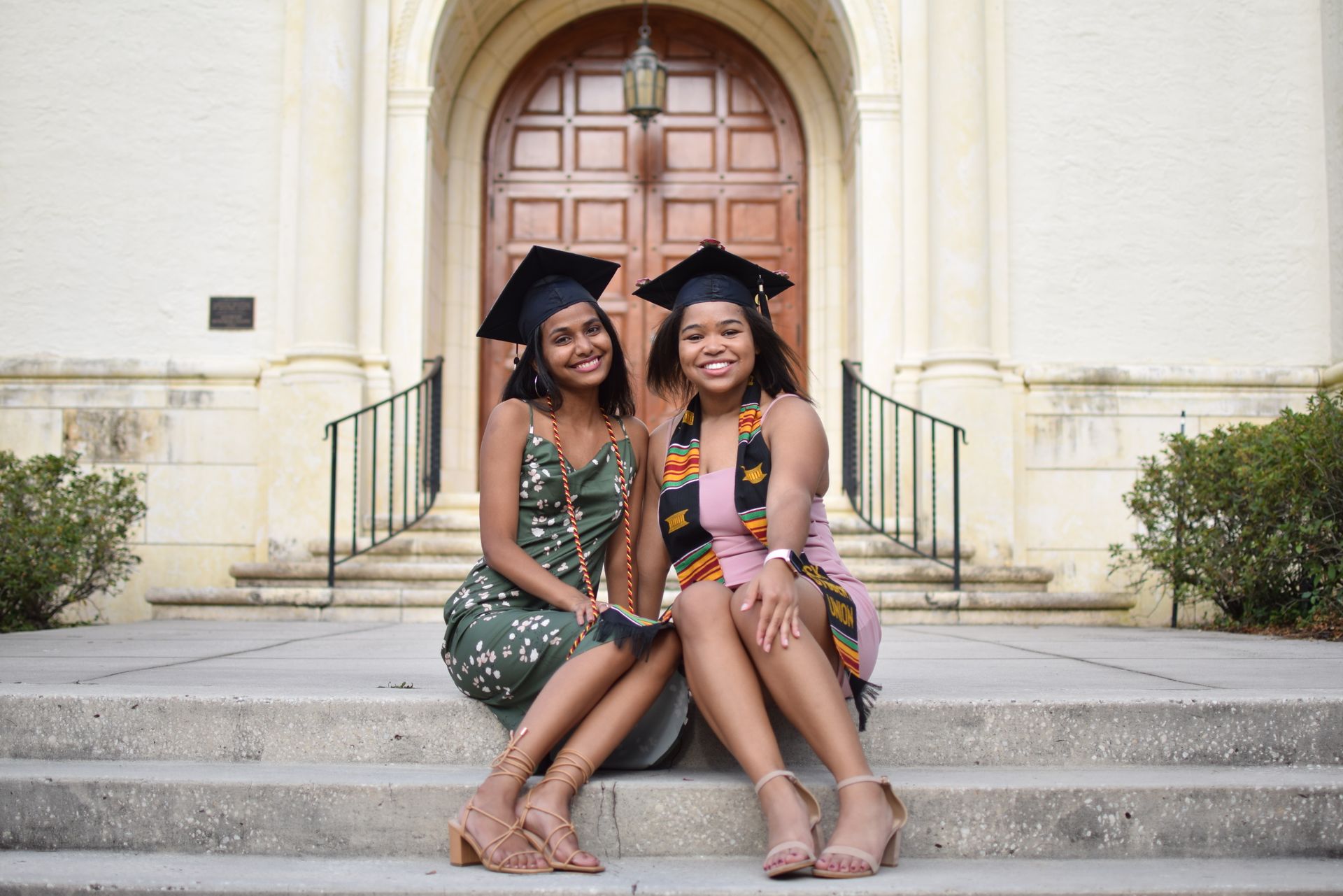 Two smiling graduates in caps and gowns sitting on steps outside a building.