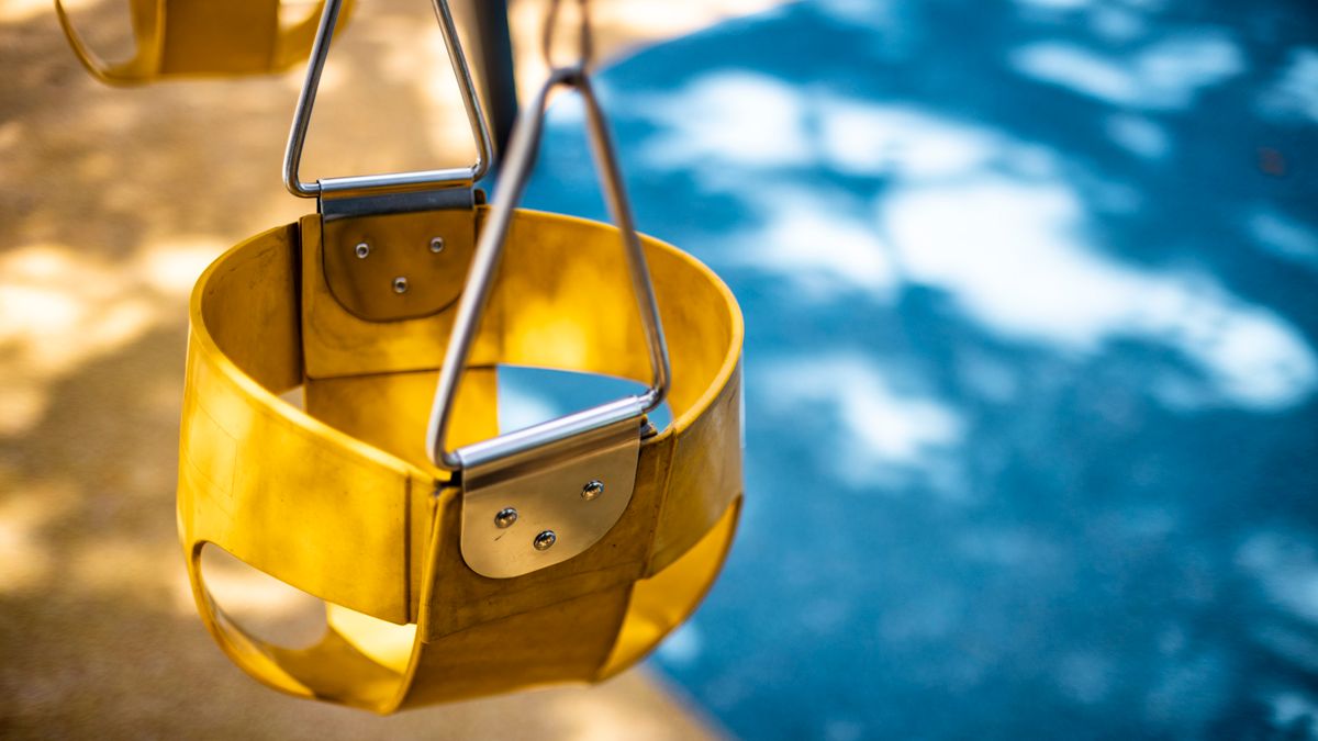 Close-up of a yellow playground swing seat in a sunlit park setting, casting shadows on the pavement.