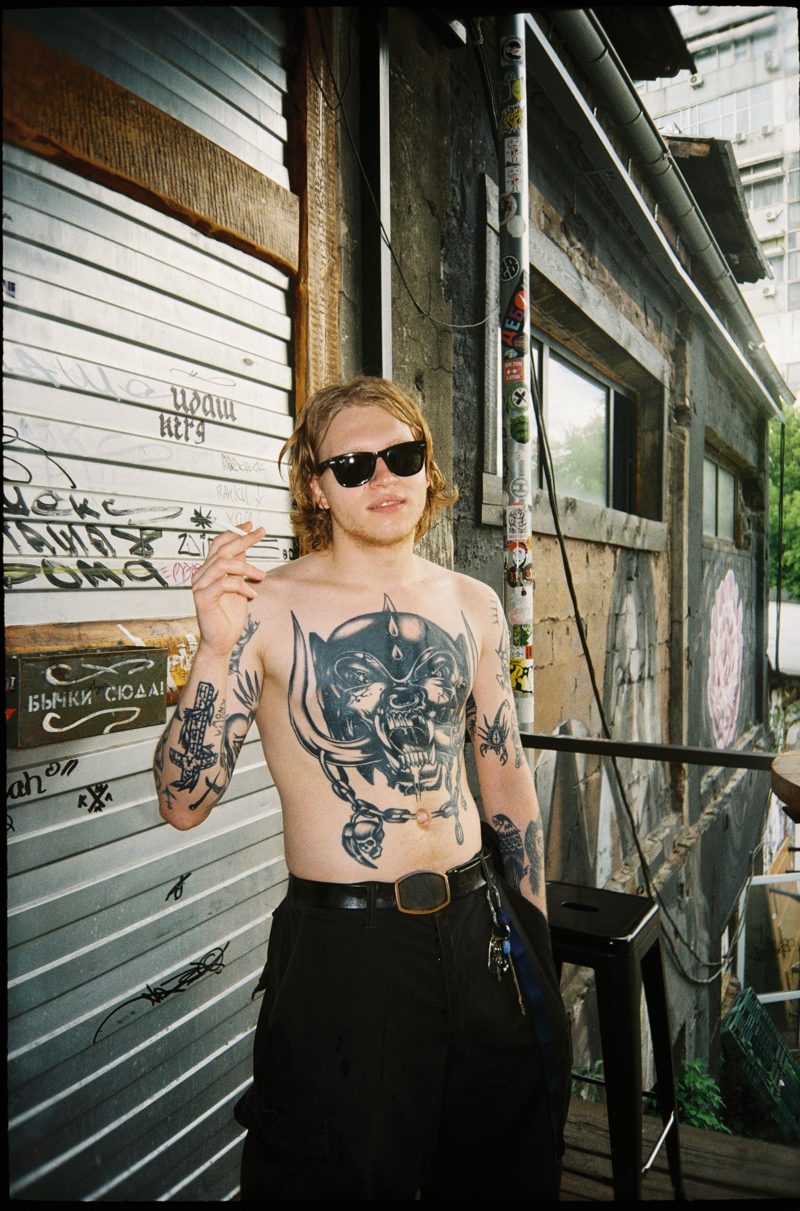 Shirtless man with tattoos and sunglasses smoking outside in Moscow street.