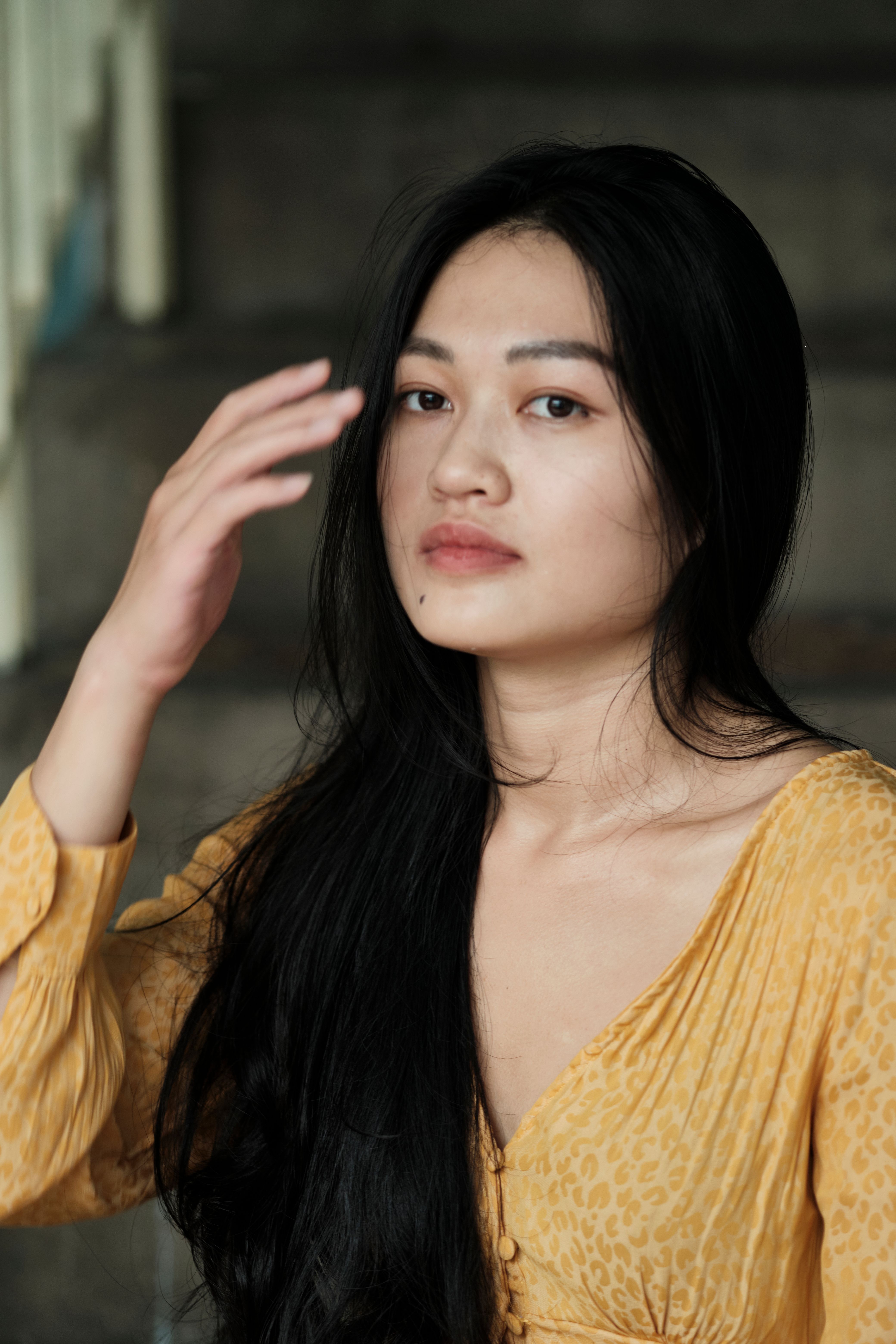 A close-up portrait of an East Asian woman indoors, wearing a yellow dress, with a soft and pensive expression.