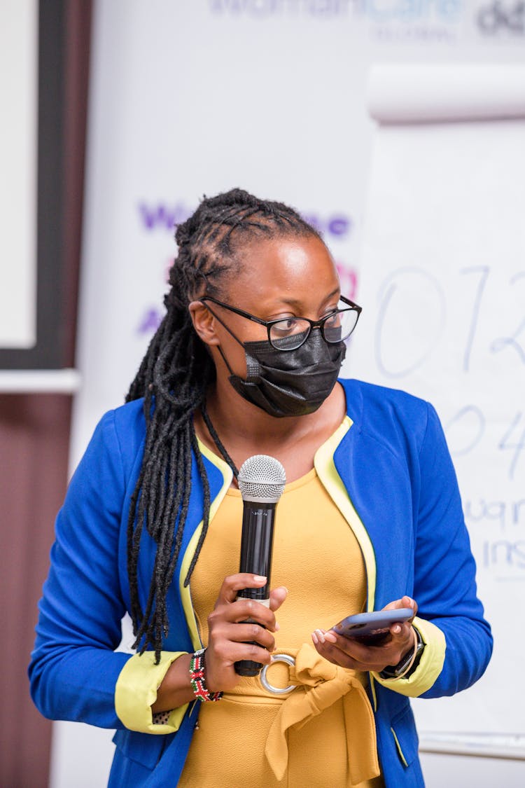 Woman Holding A Microphone While Wearing A Black Facemask 