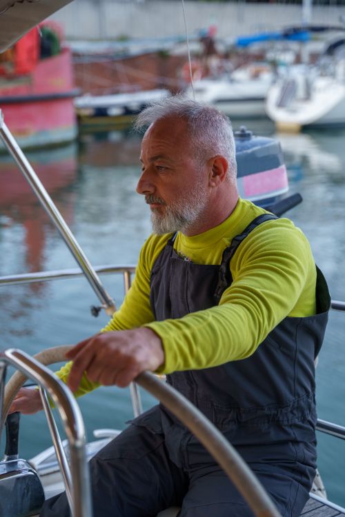 Free Mature man with gray hair steering a boat in a calm marina setting, focused and serious. Stock Photo