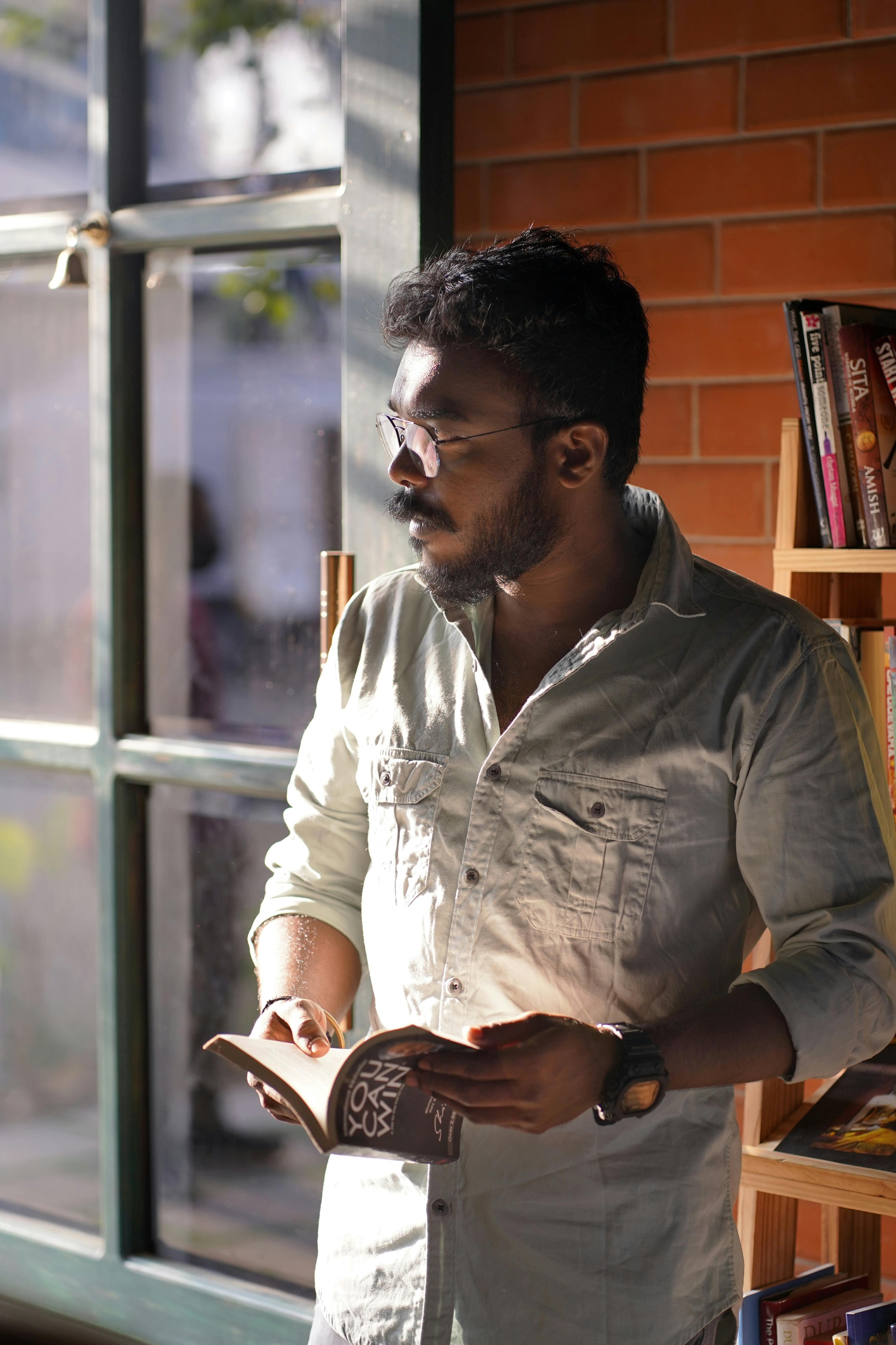 South Asian man reading a book by the window in a cozy library setting.
