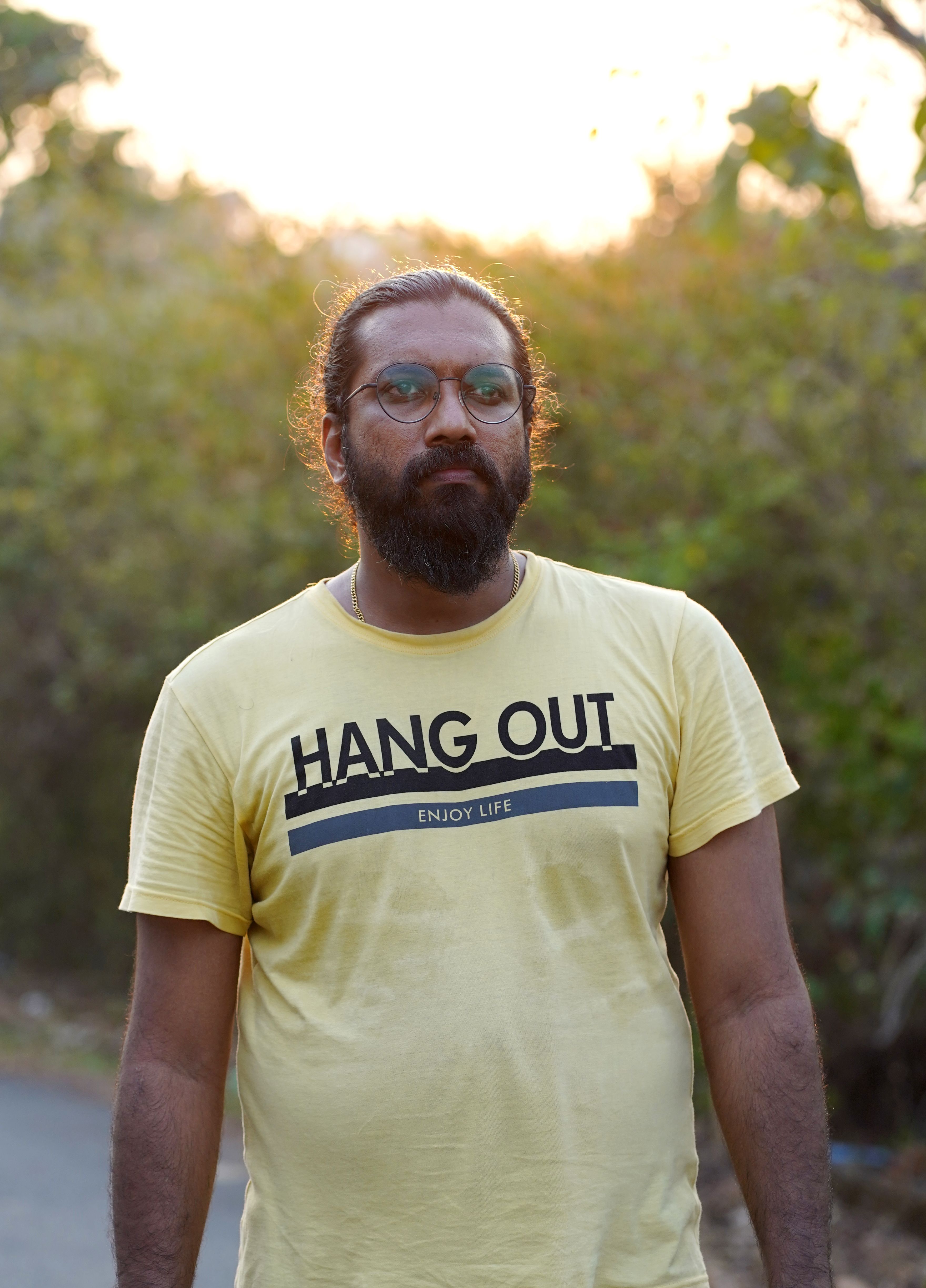 Man wearing yellow 'HANG OUT' t-shirt standing outdoors in Kerala with natural background.