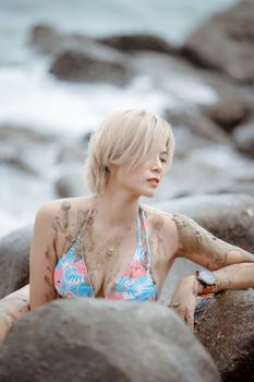 Blonde woman in floral bikini poses peacefully amid coastal rocks and muddy water.