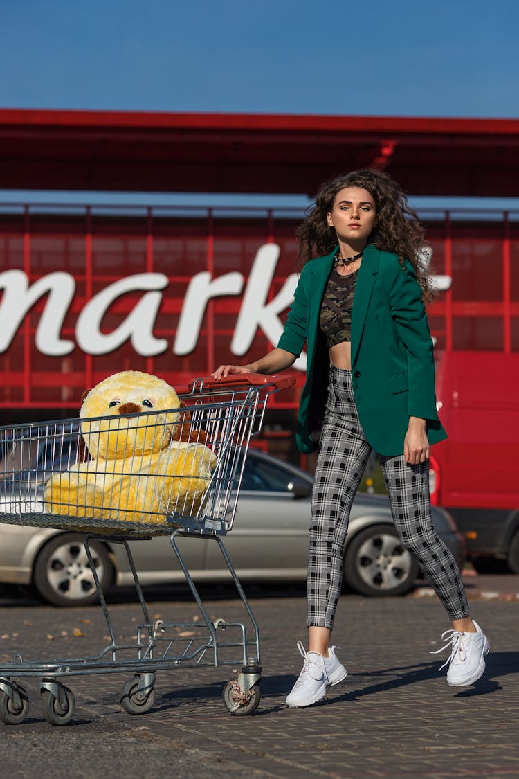 Woman Wearing Checked Trousers Walking Outside A Supermarket With A Toy In A Trolley