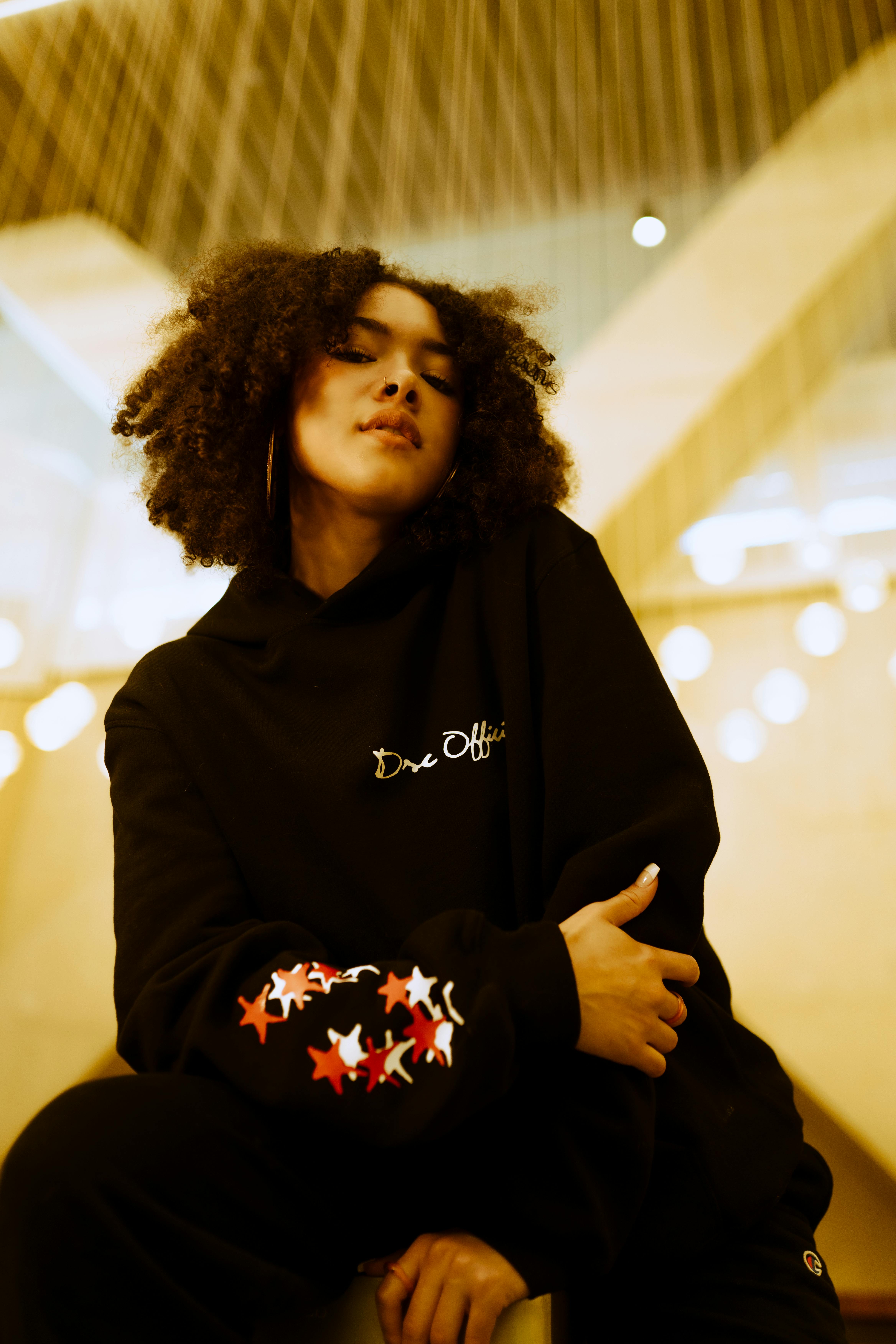 A fashionable woman with afro hair poses indoors under warm lighting.