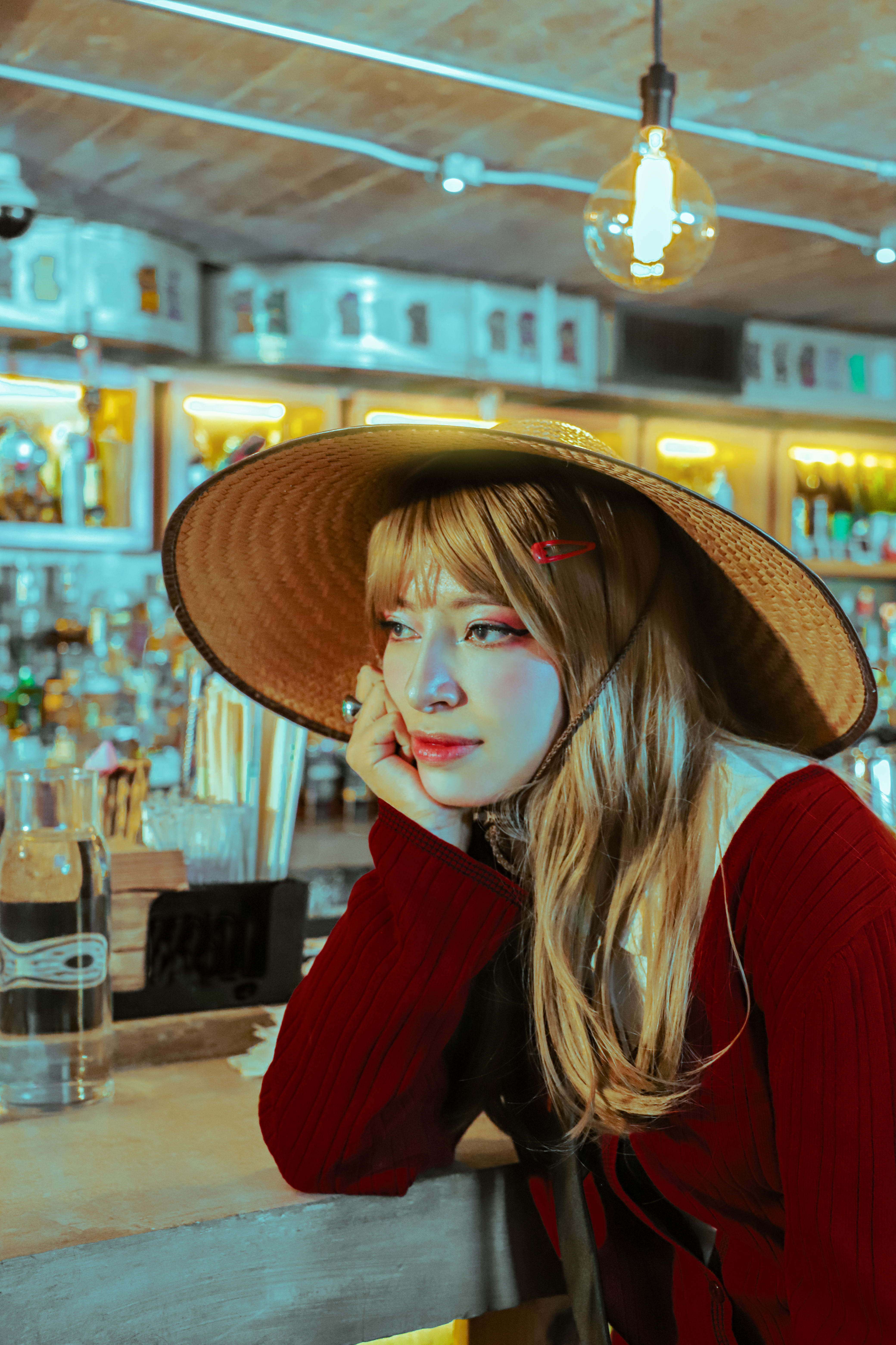 A woman in a red blouse and straw hat leans on a bar counter, gazing thoughtfully.