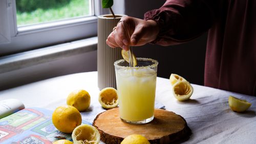 Free A glass of freshly squeezed lemonade with lemons on a wooden board by a window. Stock Photo