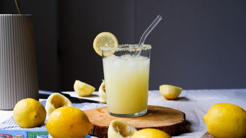 Free Fresh lemonade in a glass with a straw, surrounded by lemons on a wooden board. Perfect summer refreshment. Stock Photo