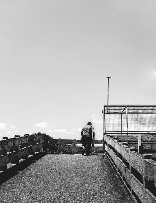 A lone man walks on a concrete bridge under a vast sky, captured in classic black and white.