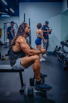 Muscular man sitting on a weight bench in a modern gym, surrounded by fitness enthusiasts.