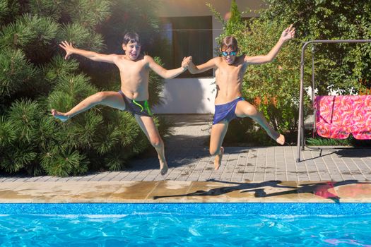 Two boys joyfully jumping hand-in-hand into a bright outdoor swimming pool during summer.