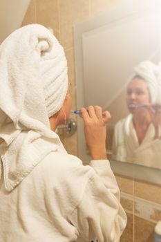 A woman in a bathrobe brushes her teeth while looking in the bathroom mirror, promoting daily hygiene.
