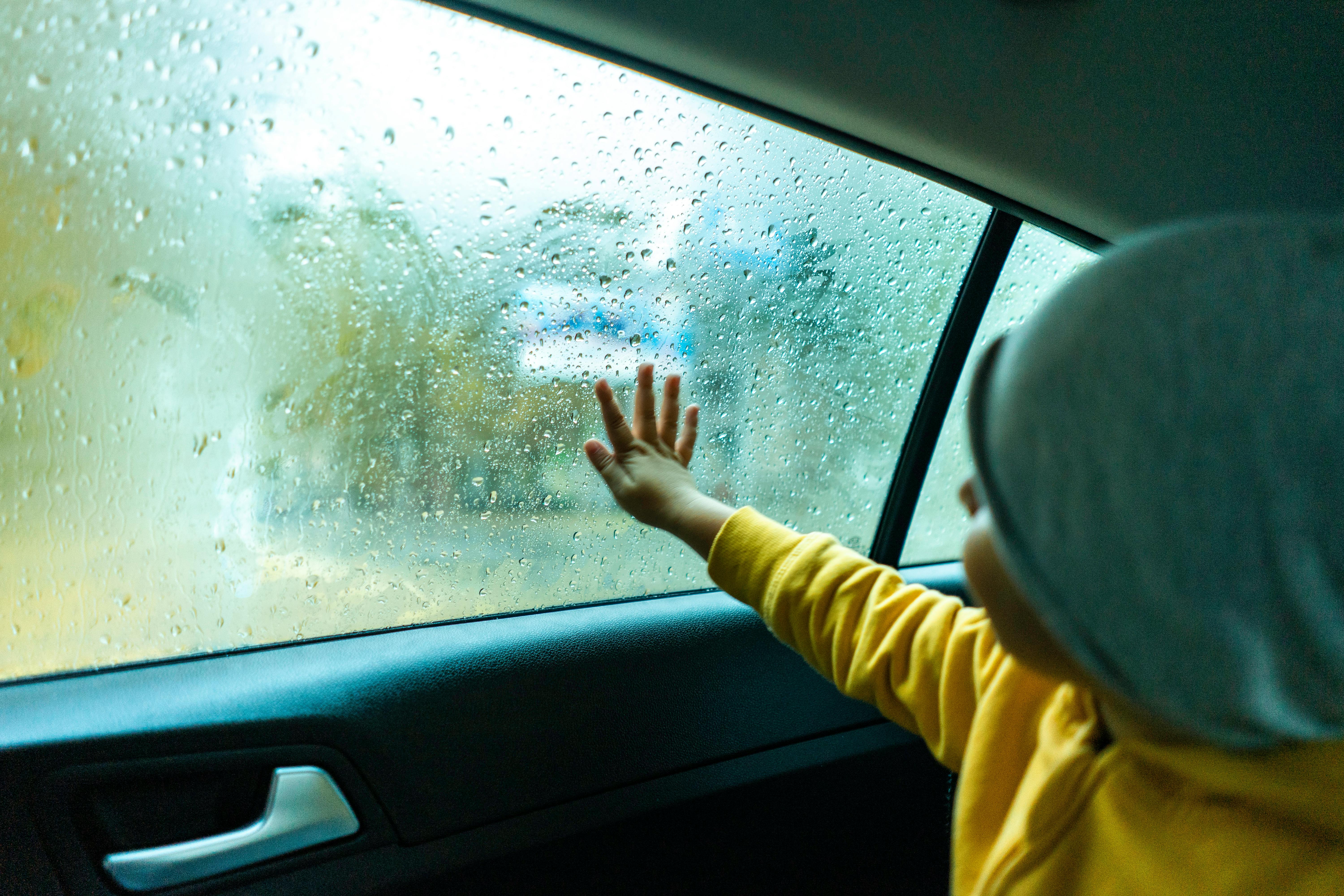 Child touching car window with raindrops, conveying a sense of curiosity and wonder during a rainy drive.
