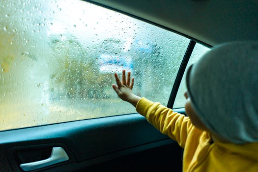 Child touching car window with raindrops, conveying a sense of curiosity and wonder during a rainy drive.
