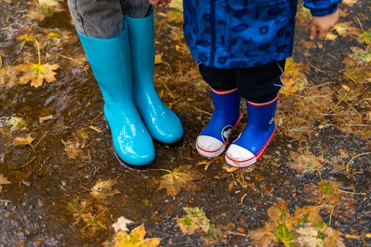 Two children wearing bright rubber boots splash in autumn puddles covered with fallen leaves.