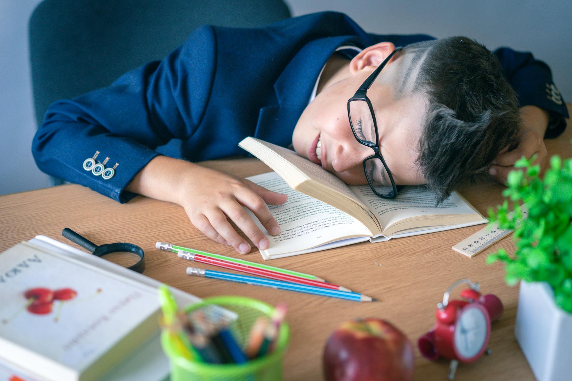 A tired schoolboy in glasses sleeping on a desk surrounded by study materials.