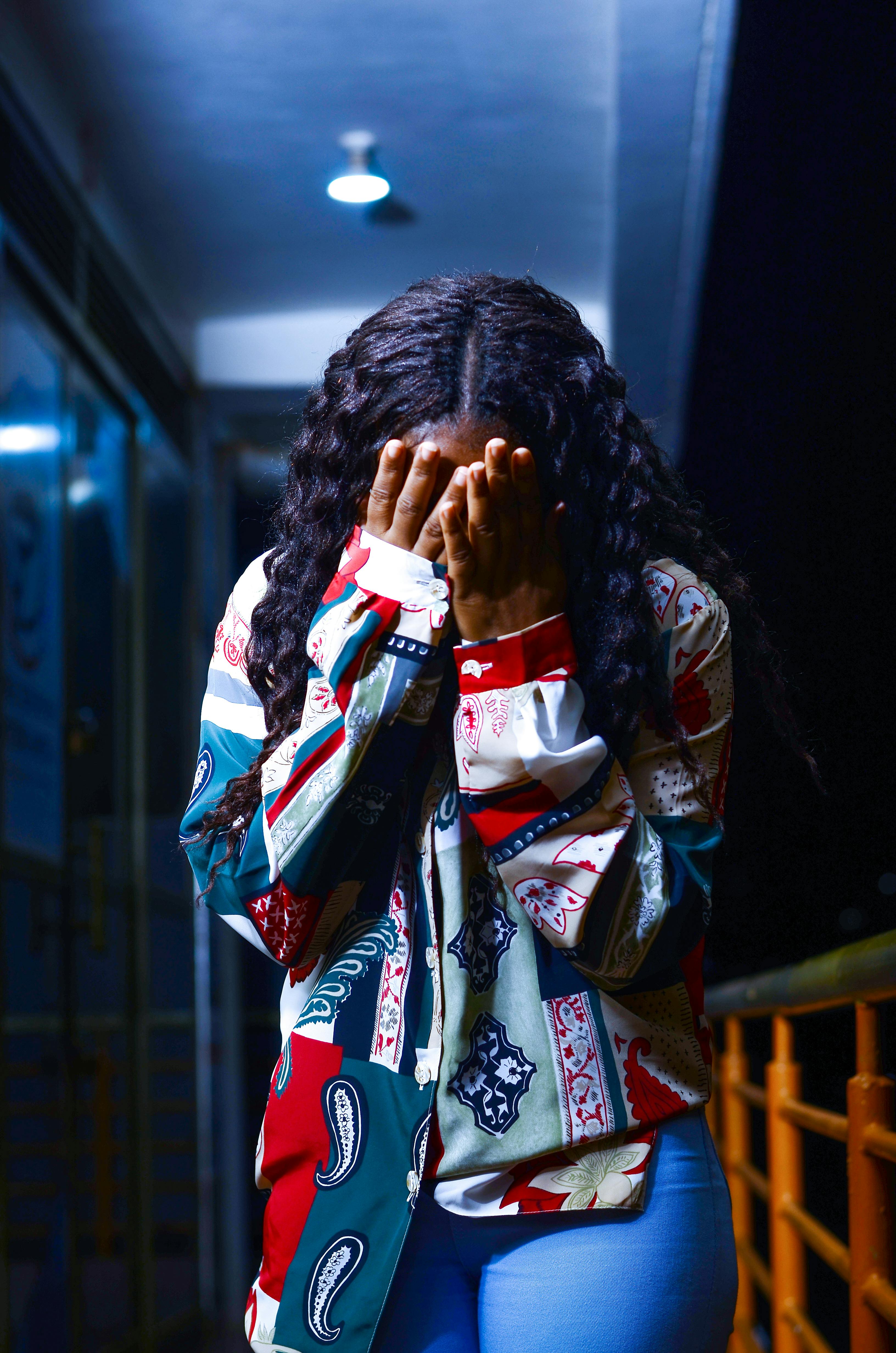 A woman covers her face with her hands at night, wearing a colorful patterned shirt.