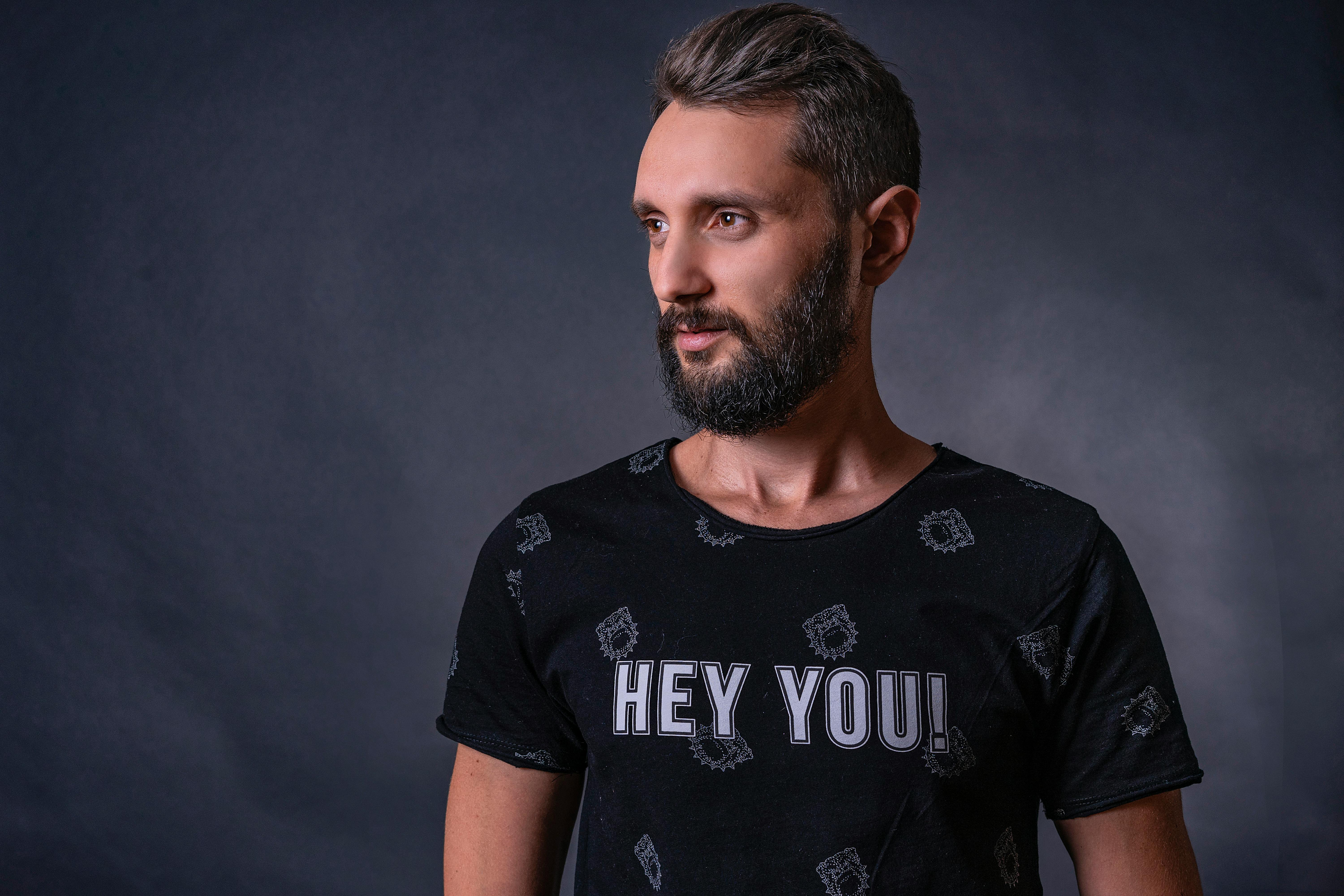 Adult man with graying hair and beard in a studio setting, wearing a 'Hey You!' t-shirt.