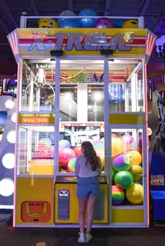 A young woman playing with a colorful claw machine at an arcade, filled with vibrant balls and neon lights.