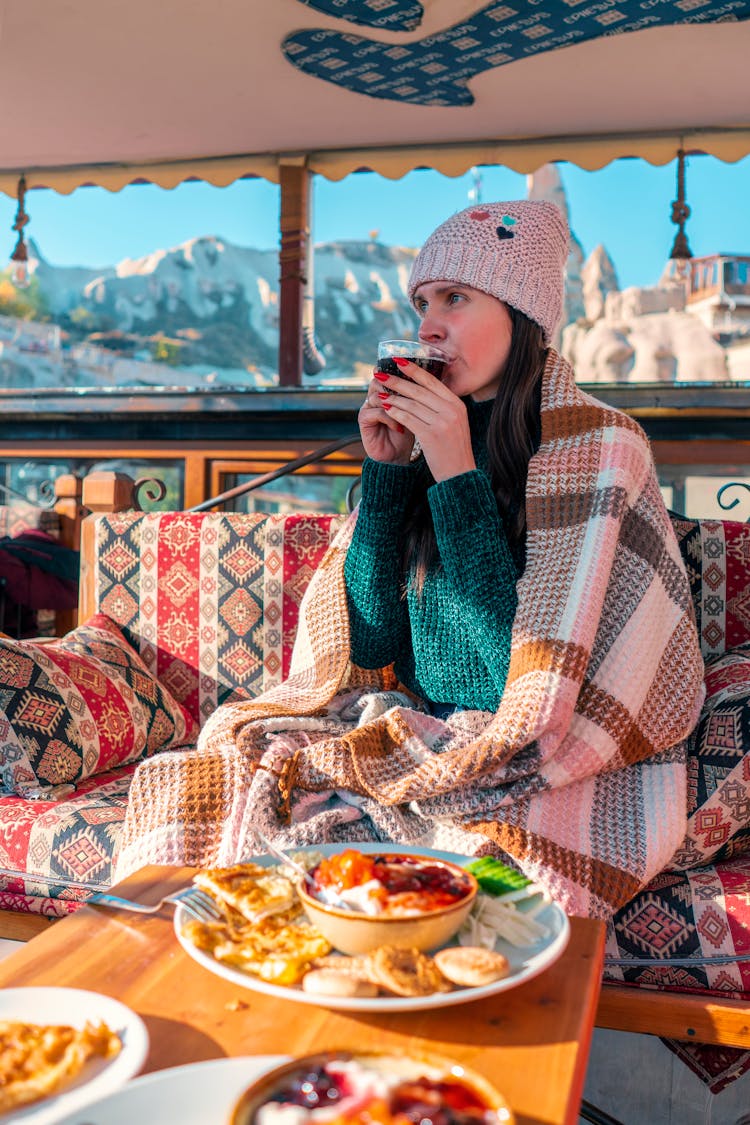 Woman Wearing Winter Hat And Wrapped In A Blanket, Enjoying Meal In A Mountain Village