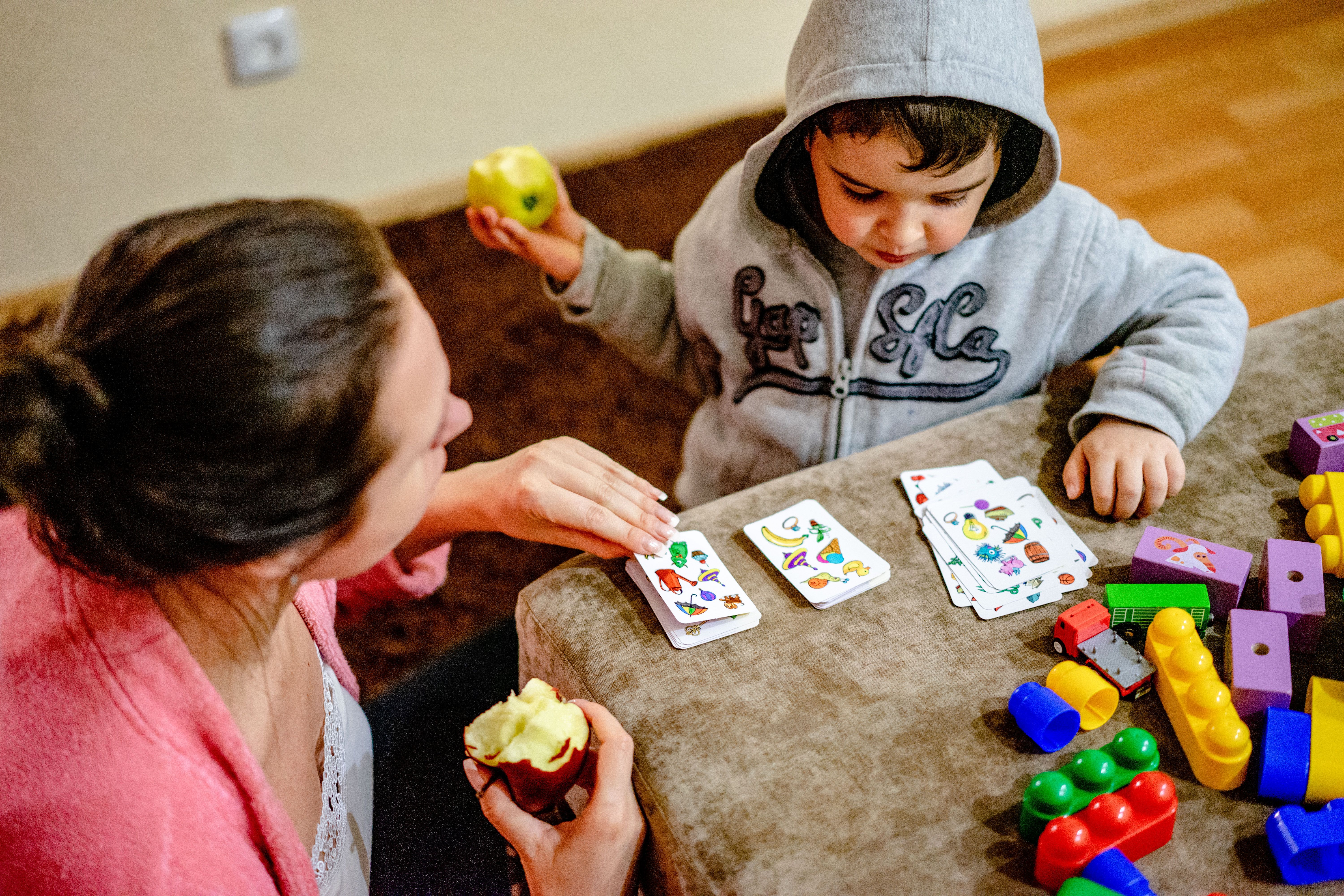 Mother and child playing a card game surrounded by colorful toys indoors.