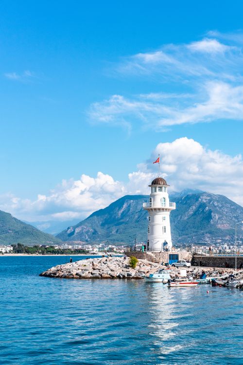 Free Beautiful seaside view of a lighthouse against Alanya's mountainous landscape and blue sky. Stock Photo