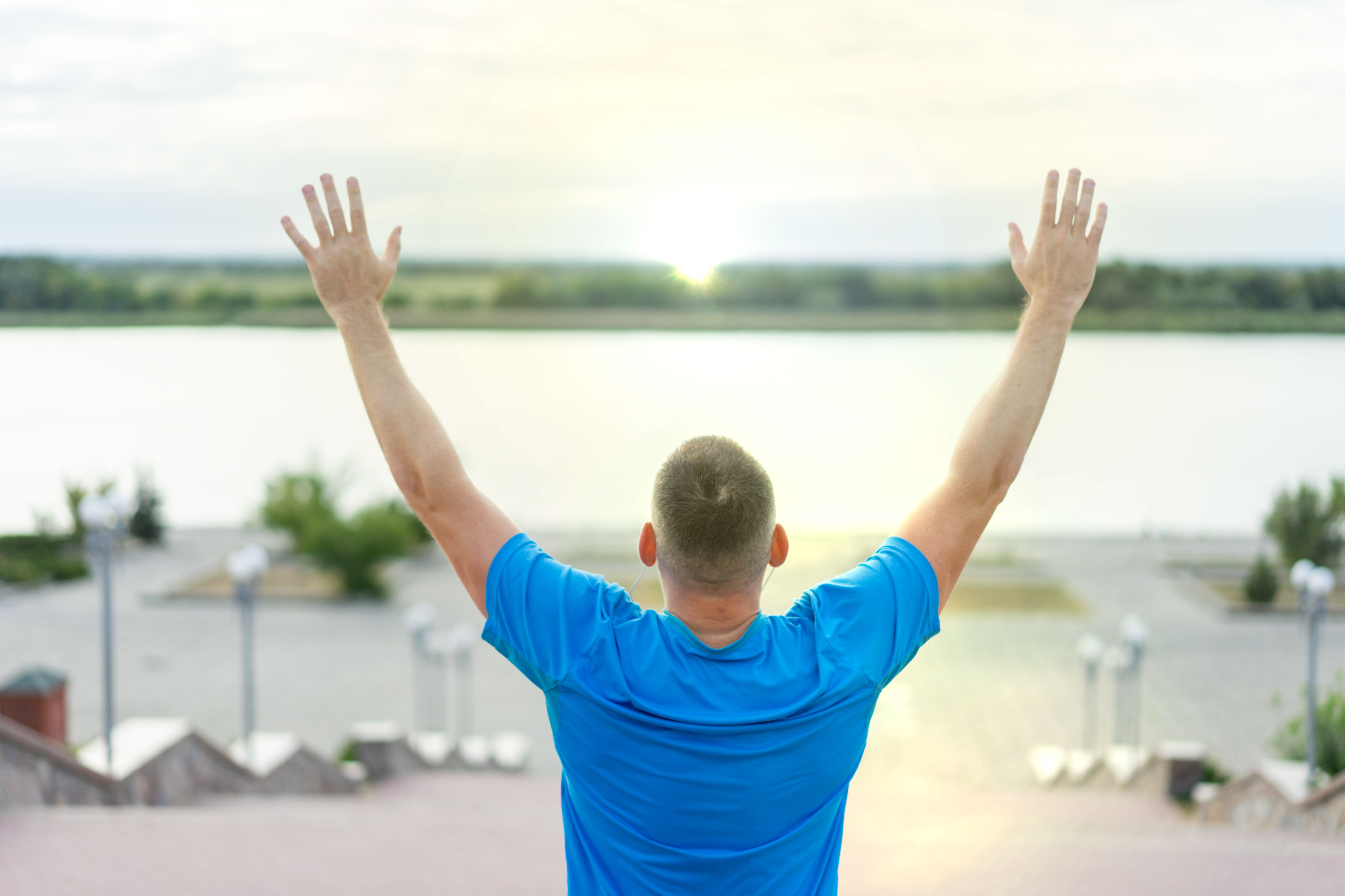 A person in a blue shirt standing with arms raised, facing a river at sunrise.