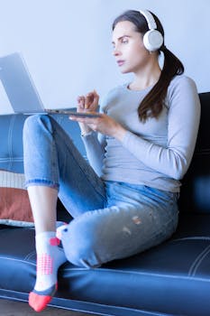 Woman in casual attire working on a laptop with headphones, relaxed on a sofa.