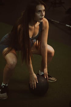Focused woman in sportswear crouching with medicine ball in gym setting.