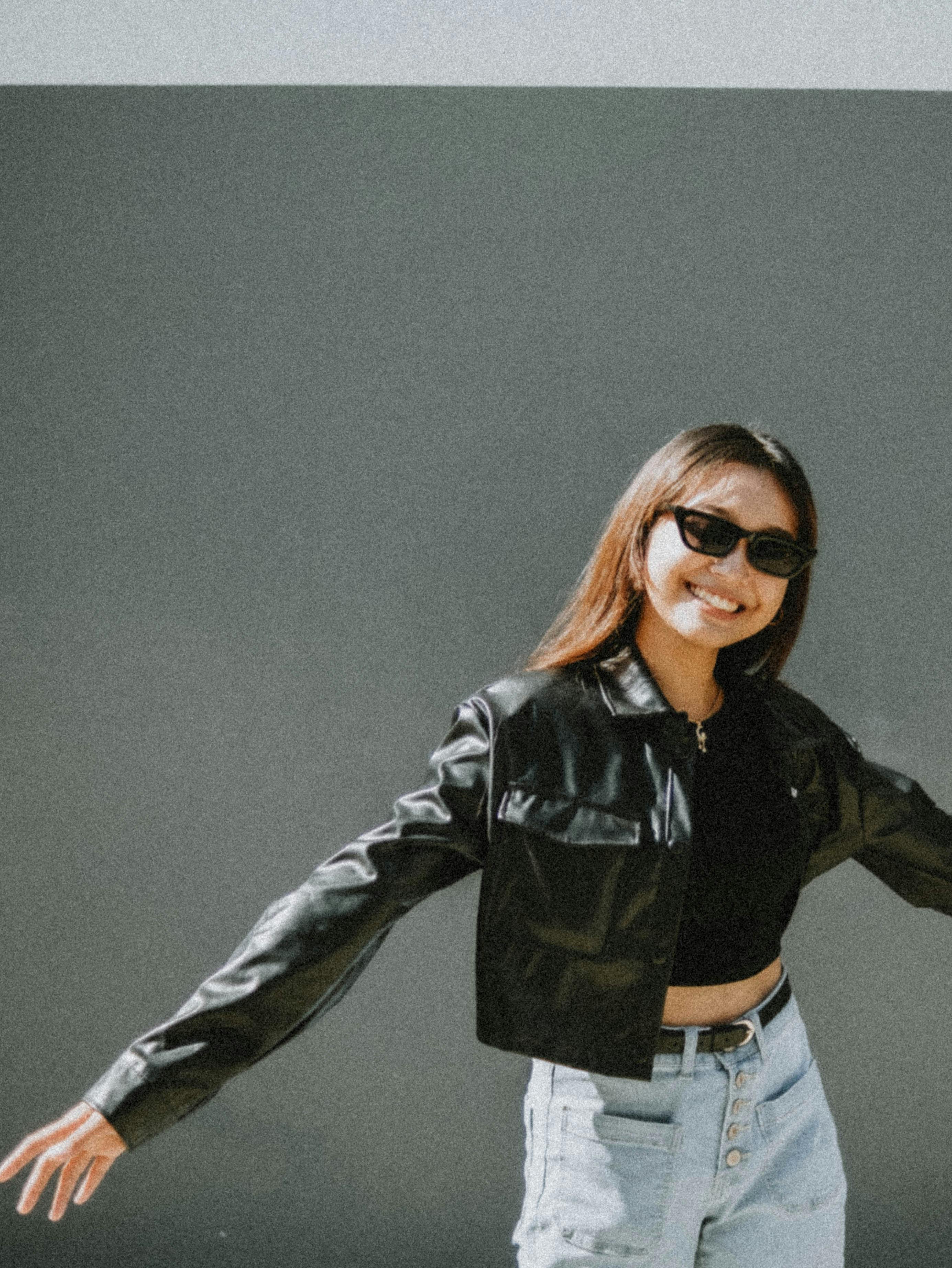 Stylish young woman in sunglasses and black leather jacket posing confidently outdoors.
