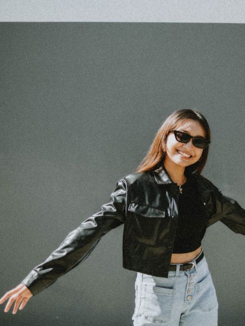 Stylish young woman in sunglasses and black leather jacket posing confidently outdoors.