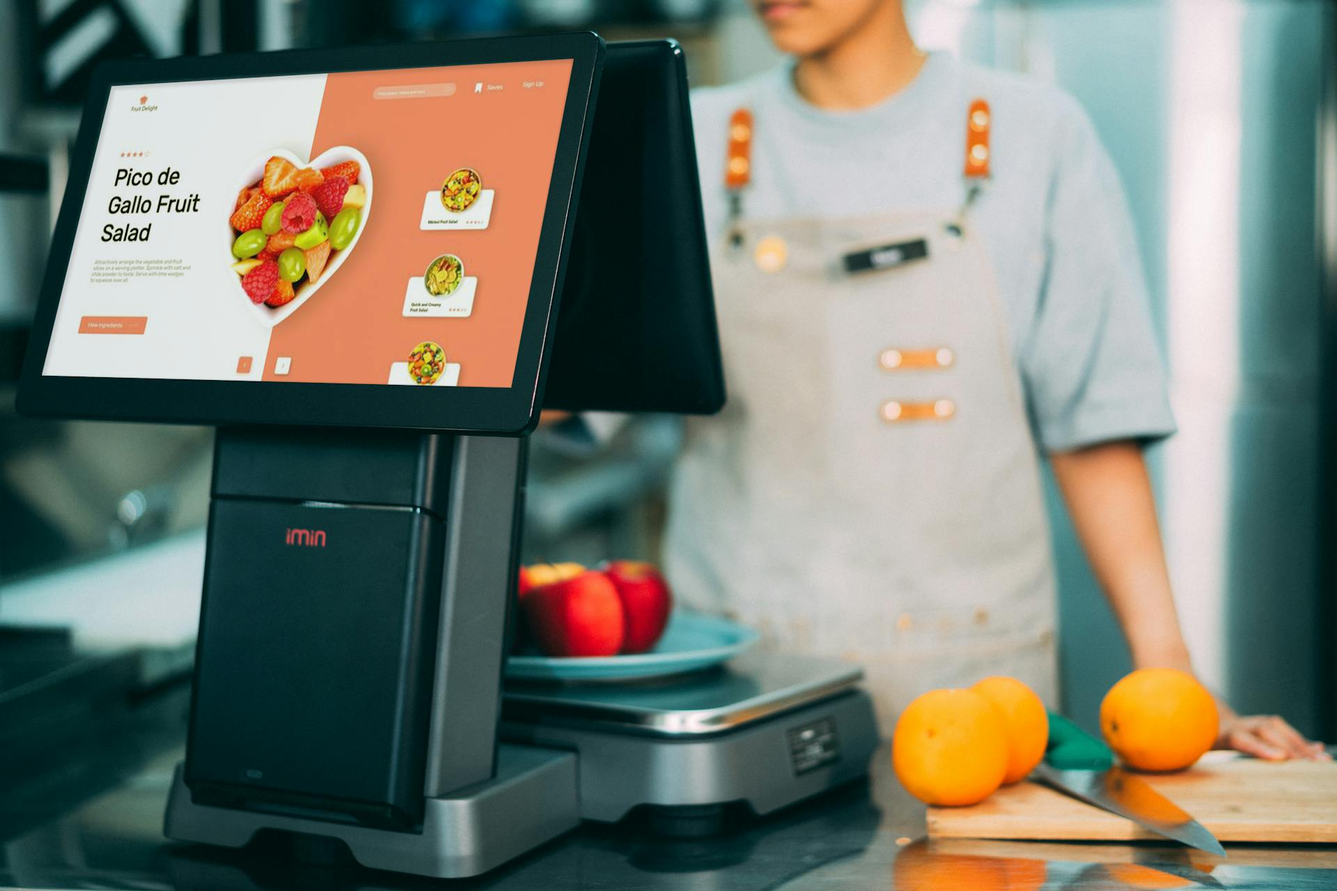 Digital checkout system in use at a grocery store with fresh fruits and a cashier in sight.