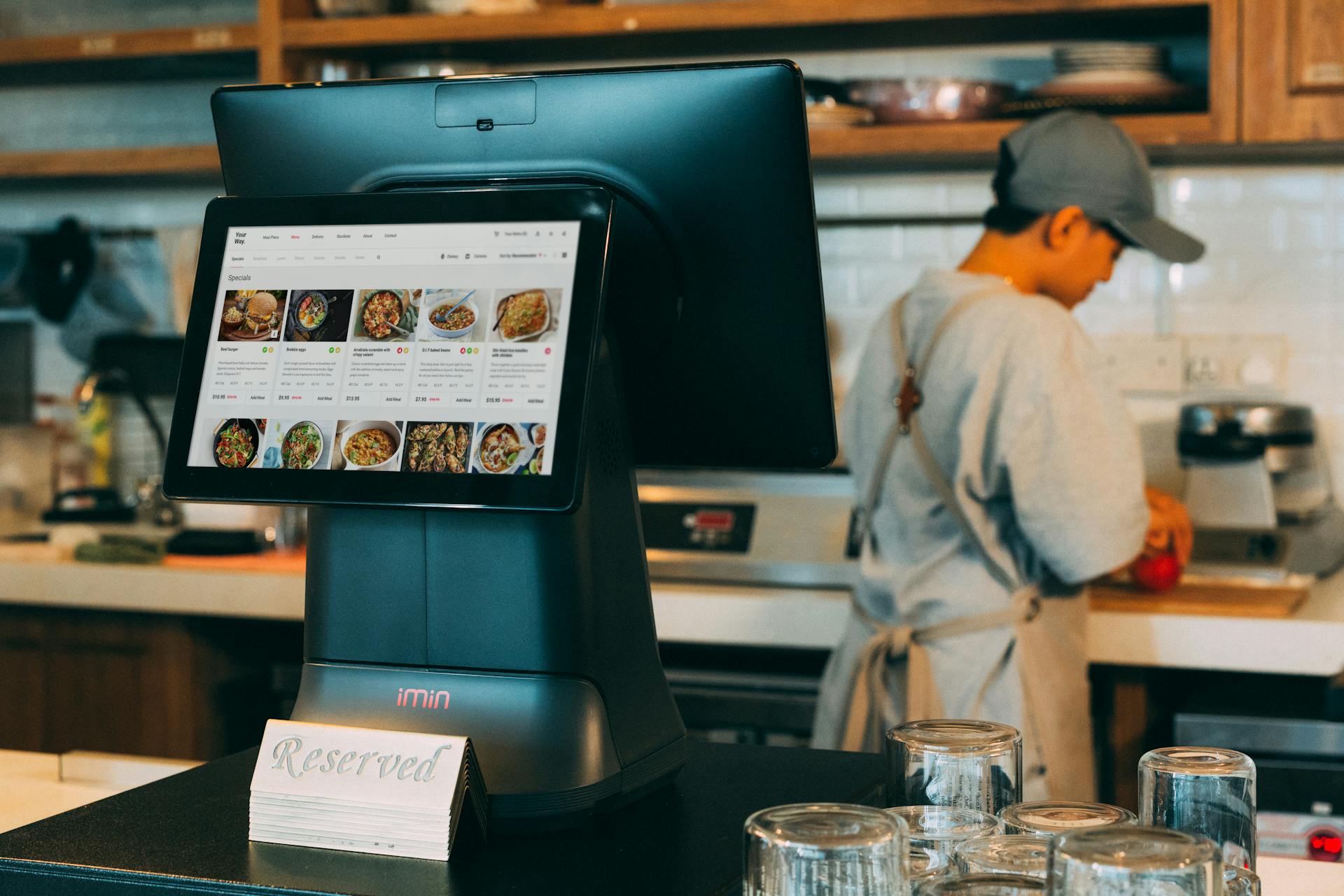 Digital ordering system in a restaurant kitchen with a touchscreen display and a chef at work.