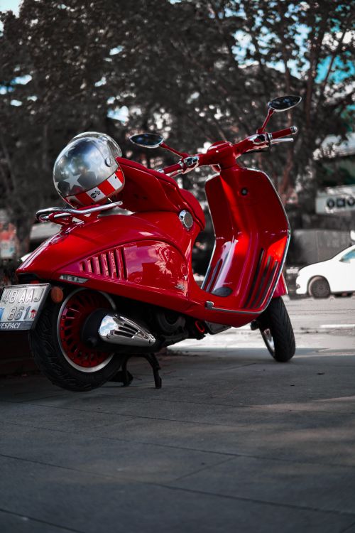 Free A striking red scooter parked on a city sidewalk with trees in the background. Stock Photo