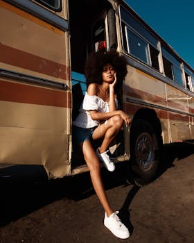 Fashion model with afro hairstyle posing next to a vintage bus under bright sunlight.