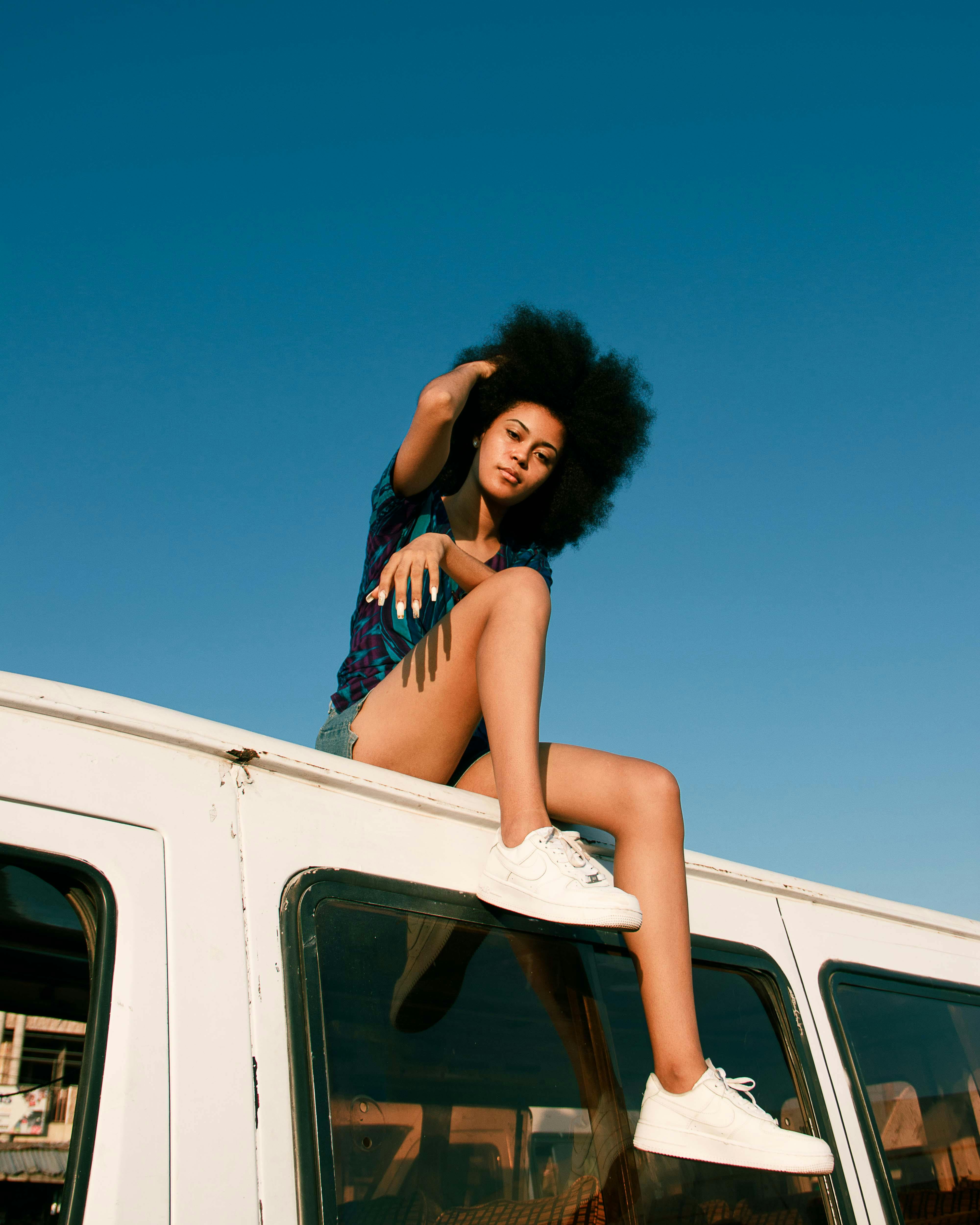 A young woman with afro hair sits on a van roof against a bright blue sky, embodying a carefree and vibrant lifestyle.