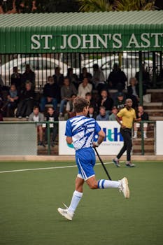 A soccer player in action during a competitive match at St. John's Astroturf, with audience in the background.