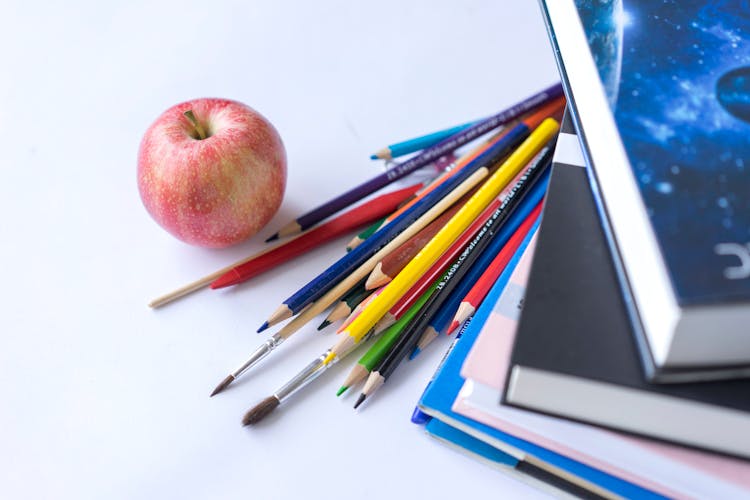 Apple Fruit Near Color Pencils Paint Brush And Books On White Surface