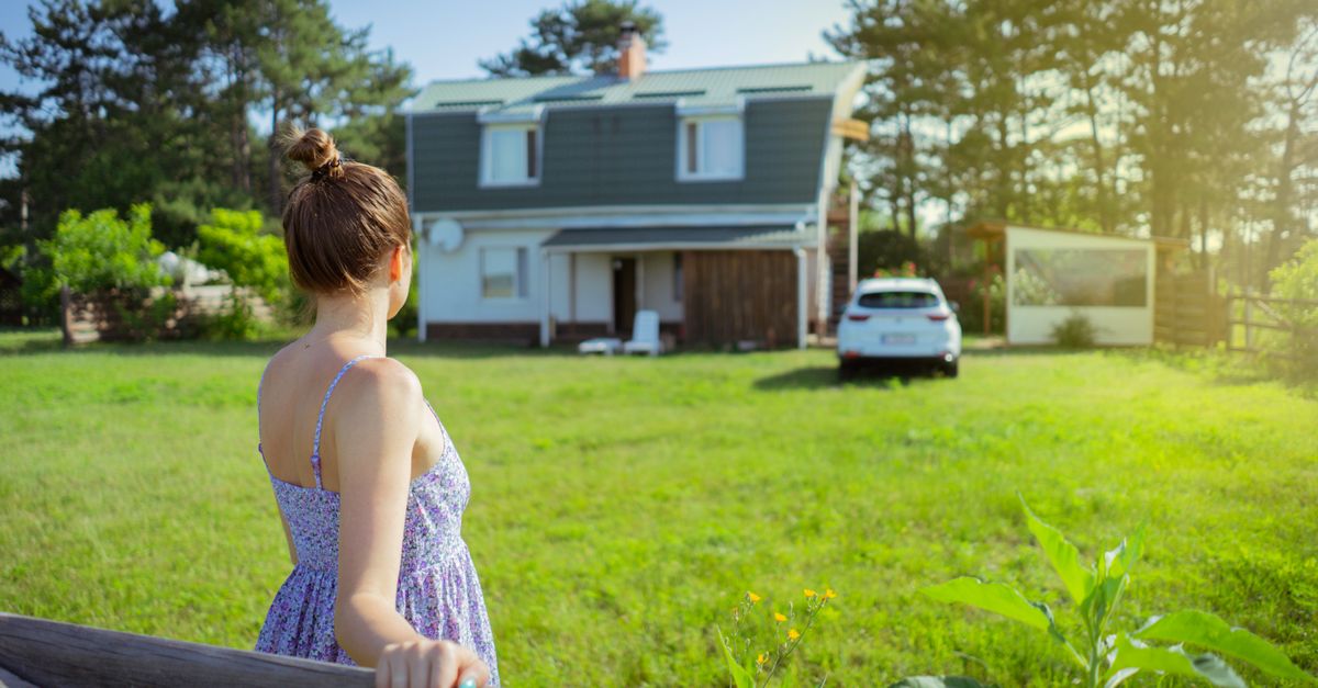 A woman in a sundress stands by a fence admiring a countryside house on a sunny day.