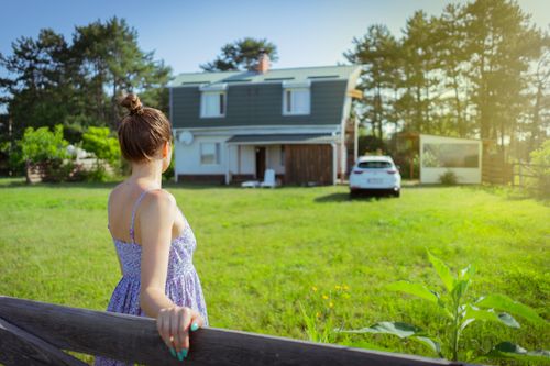 Free A woman in a sundress stands by a fence admiring a countryside house on a sunny day. Stock Photo
