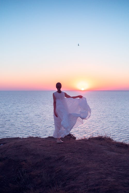 A Woman in White Dress Standing Near Body of Water during Sunset