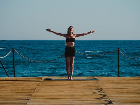 A woman in activewear stands in a yoga pose on a wooden dock near the sea, embracing a tranquil moment.