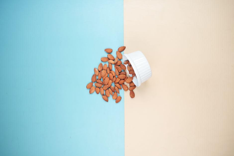Hạt Hạnh Nhân: "Siêu Thực Phẩm" Cho Làn Da & Vóc Dáng – Cẩm Nang 2026 4 A bowl of almonds spills onto a blue and beige backdrop. Minimalist food concept.
