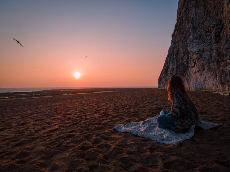 Woman Sitting On A Blanket On Sand Watching Sunset