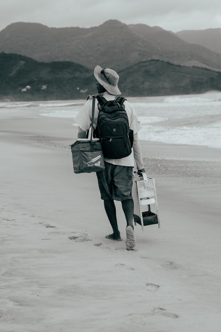 Grayscale Of A Man In White Polo Shirt Walking On Seashore With Black Backpack Bag