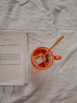 Flat lay of an open book and a mug with spoon on a white fabric background, depicting a cozy reading moment.