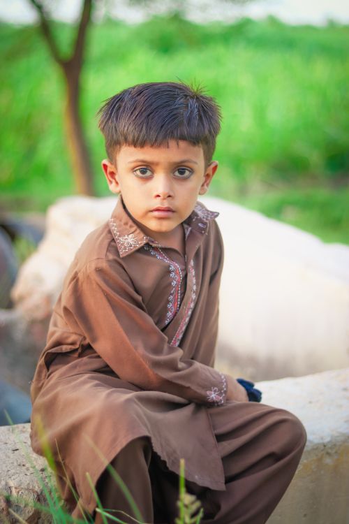 Free A young boy in traditional clothing sits outdoors, looking directly at the camera. Stock Photo